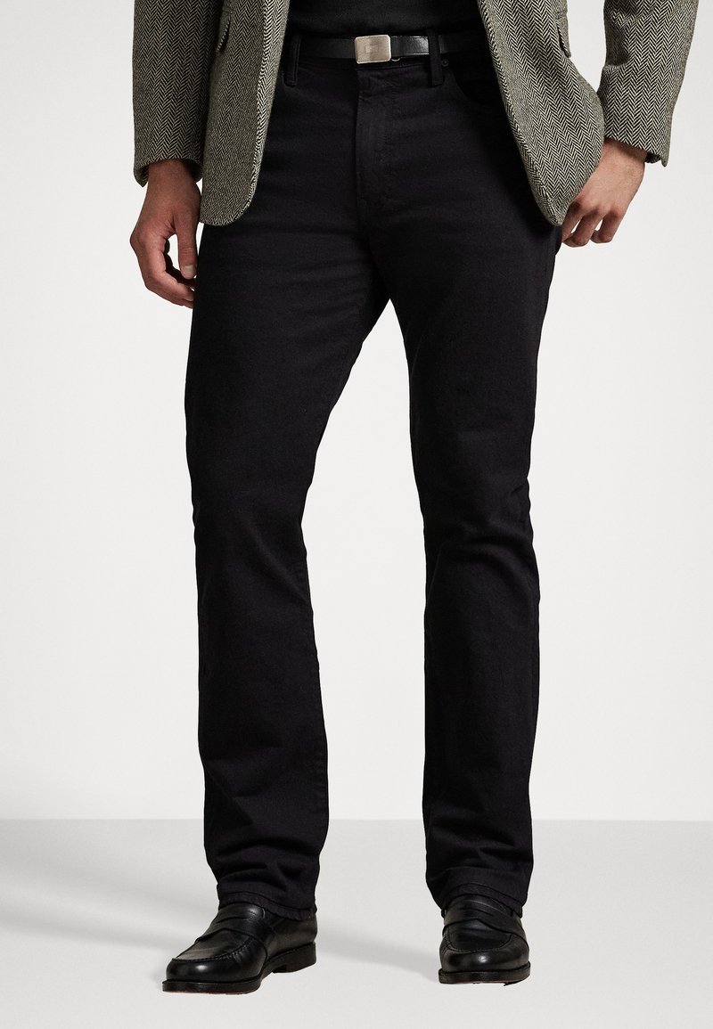 Jeans pretos de perna reta com uma textura suave, usados com uma camisa escura e um blazer de padrão espinha de peixe, apresentando uma fivela de cinto prata e sapatos pretos.