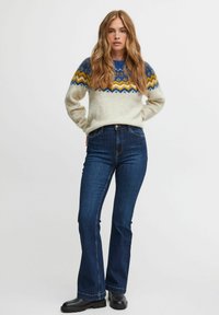 Jersey de punto en crema con patrones geométricos en azul y amarillo, combinado con jeans acampanados de color azul oscuro y botas negras de tobillo.