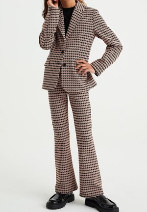 Brąsu i czarne ubranie w wzór houndstooth, składające się z dopasowanej marynarki i spodni w dzwony, wykonane z teksturowanej tkaniny. Całość dopełniają masywne czarne buty.