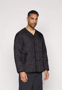 Black quilted jacket with a deep V-neck, front buttons, and side pockets. Textured fabric with subtle wave patterns, paired with a beige shirt underneath.