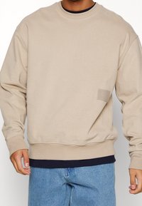 Beige crewneck sweatshirt van zachte stof, met ribbelmanchetten en -zoom, en een klein logo-label aan de voorkant nabij de zoom.