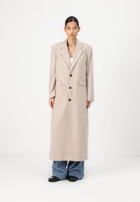 Long beige striped coat with large lapels and five buttons. The fabric appears textured, paired with loose blue jeans and black sandals.