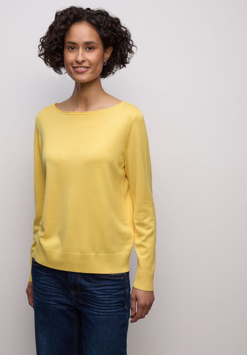 Pull jaune tricoté avec un large col et des manches longues, associé à un jean bleu foncé. Texture lisse, coupe décontractée.