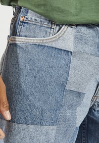 Denim jeans featuring a patchwork design in varying shades of blue, with visible stitching and copper hardware details at the waist.