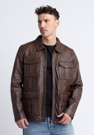 Mann trägt dunkelbraune Lederjacke mit vier geknöpften Taschen über schwarzem Hemd und blauen Jeans, schaut zur Seite.