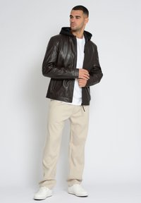 Brun læderjakke med sort hætte, lynlåslukning og to sidelommer. Båret over en hvid t-shirt og crèmefarvede bukser.