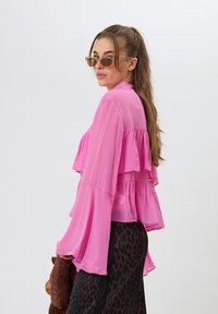 Vaaleanpunainen sifonkipaita, jossa on poimutettuja kerroksia ja kellohelmaiset hihat, yhdistetty tumman leopardikuosiset hameen kanssa. Asusteina kevyet ruskeat aurinkolasit.