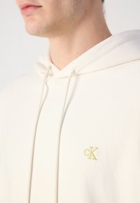 Felpa crema realizzata in morbido tessuto, caratterizzata da una tasca a marsupio frontale e da un logo "cK" ricamato in neon giallo sul petto.