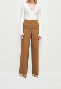 Pantalon marron à jambes larges avec une taille haute, fabriqué en tissu lisse. Associé à un haut blanc ajusté à manches longues avec un décolleté en V profond.
