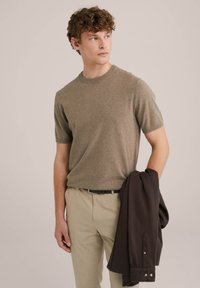 Short-sleeve, round-neck shirt in a light brown melange knit, paired with beige trousers. A dark brown jacket is draped over one arm.