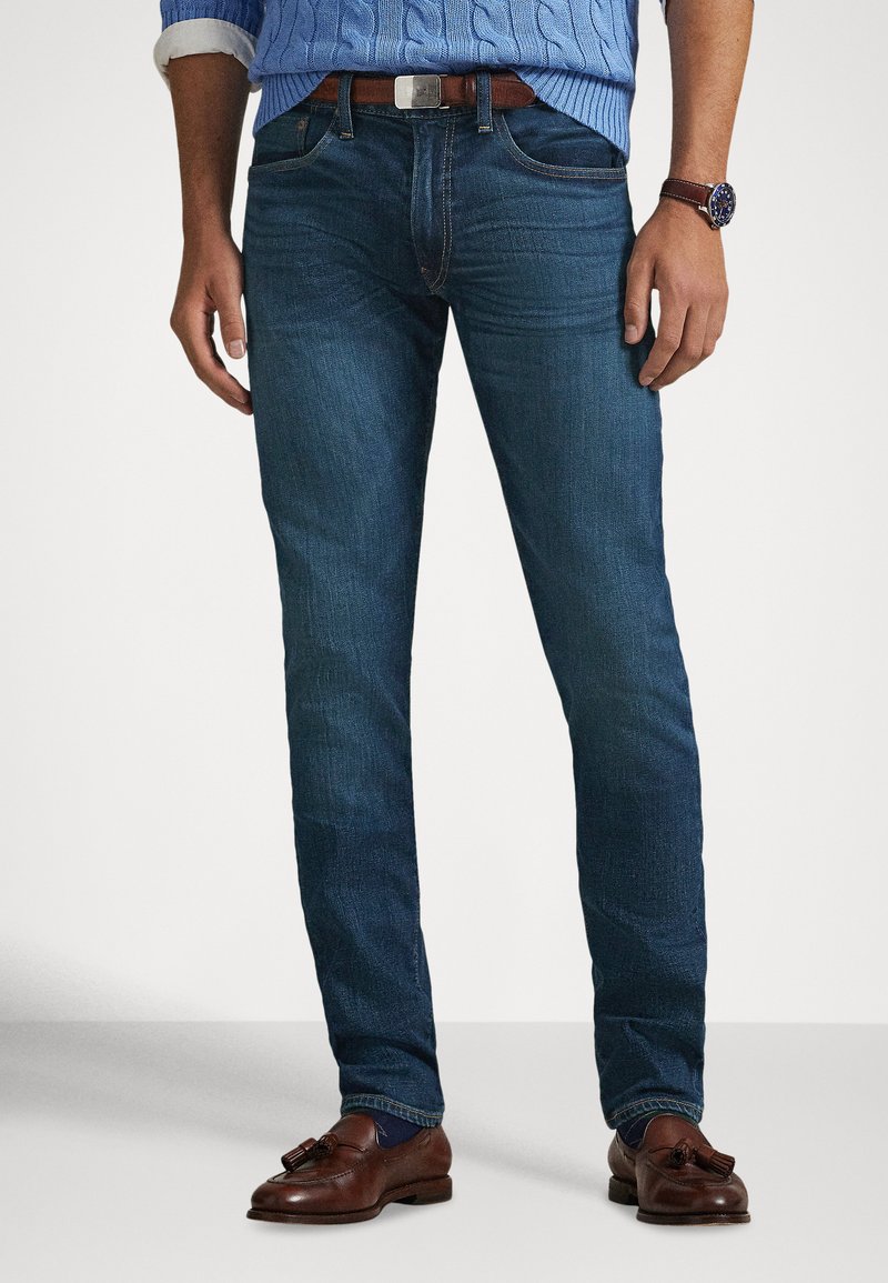 Jeans de ganga azul escuro com corte slim, com bolsos frontais e um cinto de couro castanho. O tecido tem uma textura suave e um desbotamento subtil.