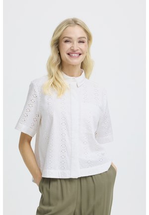 Naeratav naine pikkade blondide juustega kannab valget lühikeste varrukatega silmapaistvat pluusi ja oliivrohelisi pükse, käed taskus, üksluisel taustal.