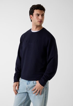 Mężczyzna w granatowej bluzie z napisem "GUESS EST. 1981", w jasnoniebieskich dżinsach, patrzący w lewo na jednolitym jasnym tle.