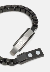 Pulsera de cadena de metal en negro, con un cierre de plata pulida que presenta el grabado "BOSS" y un conector rectangular suave con dos acentos circulares.