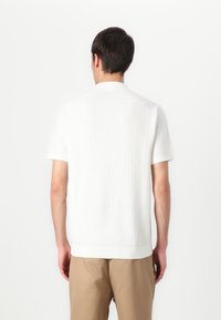 Korte ermer, hvit strikket polo skjorte med ribbet tekstur, enkel krage og rett snitt. Båret med beige bukser, viser bakfra.