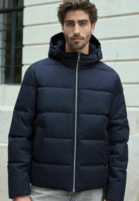 Jachetă pufă navy cu un design căptușit, închidere cu fermoar și glugă ajustabilă. Dispune de buzunare laterale și o textură de material neted.