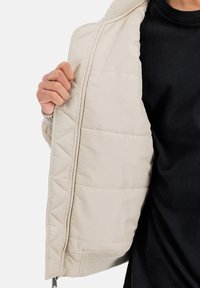 Chaquetón aislante beige con textura acolchada, cuello acanalado y abertura frontal, que revela un forro suave.