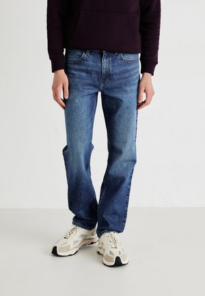 Denim jeans in medium blue with a straight leg fit, featuring a classic five-pocket design and subtle fading. Worn with beige athletic shoes.