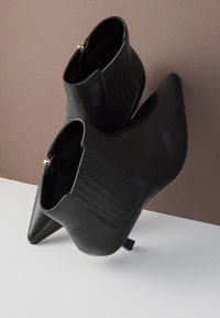 Paire de bottines en cuir noir à bouts pointus, talons bas et panneaux latéraux élastiques sur un fond bicolore marron et blanc.
