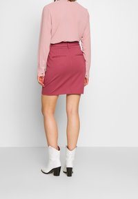 Žena v dlouhém rukávu prachaté růžové blůze, růžové sukně po kolena a bílých sáčkových botách střední výšky, stojí před jednoduše pozadím.