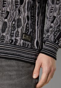 Gestrickter Pullover mit einem strukturierten Muster in Grau und Schwarz. Akzentuiert mit einem markierten Metalltag sowie gestreiften, gerippten Bündchen und Saum.