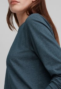 Kvinna med långt brunt hår i en blå långärmad tröja, visad från axlarna till halva överkroppen mot en enhetlig bakgrund.