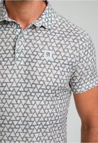 Polotröja i ljusgrått tyg med ett geometriskt mönster i blått och vitt, med krage och logotyp-patch på bröstet.