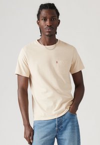Beige T-shirt lavet af bomuld, med en brystlomme der har et lille rødt logo, rund halsudskæring, og korte ærmer, parret med lyse denimjeans.