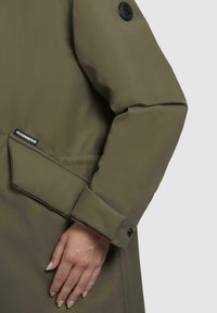 Olivengroene buitenkleding met een gladde textuur heeft een opgerold manchet, een patchlogo op de mouw en een zijzak met een zwart label.