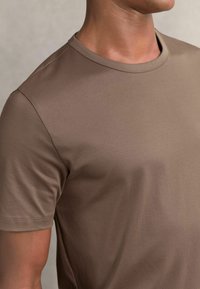Hnedé tričko s krátkym rukávom vyrobené z hladkej tkaniny, so okrúhlym výstrihom a prešitými švami, ktoré zdôrazňuje telo priliehajúci siluetu.