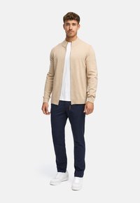 Beige zip-up cardigan over een wit T-shirt, gecombineerd met Navy broek en witte sneakers. Gladde textuur en minimalistisch ontwerp zonder patronen.