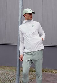 Biela šušťáková bunda so zelenými akcentmi, skombinovaná so svetlozelenými nohavicami. Bunda má kapucňu a zipsové vrecká. Čiapka je tiež svetlozelená.