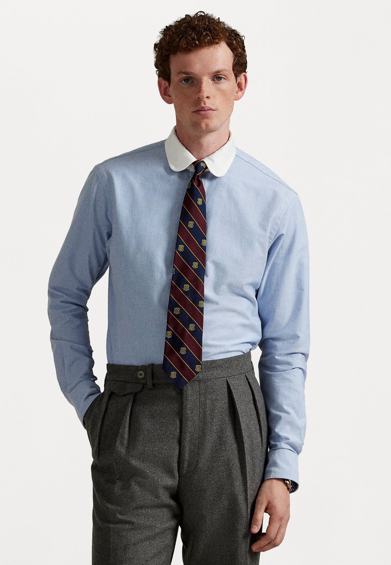 Lyseblå bomullsskjorte med hvit krage, kombinert med slips i burgunder og marine blått med gullakenter, og grå plisserte bukser.