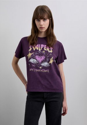 Camiseta de algodón morada que presenta una impresión gráfica con un corazón y texto, mangas cortas y un ajuste relajado. El patrón incluye estallidos y estrellas.
