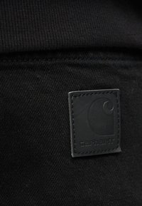 Κοντινό πλάνο μιας μαύρης ετικέτας Carhartt ραμμένης σε σκούρο ύφασμα, που αναδεικνύει την υφή και το λογότυπο της μάρκας σε casual ρούχα.