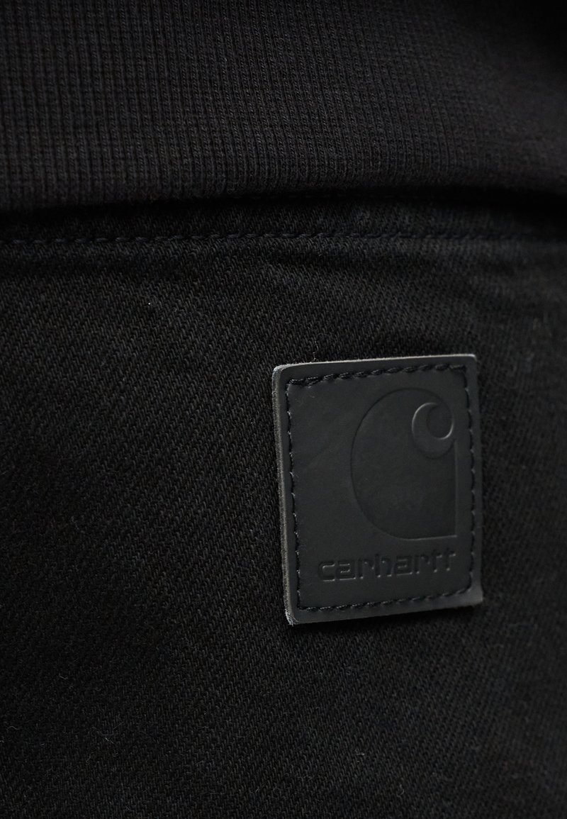 Nahaufnahme eines schwarzen Carhartt-Etiketts, das auf dunklem Stoff aufgenäht ist und die Textur sowie das Markenzeichen auf Freizeitkleidung hervorhebt.