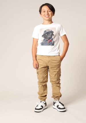 Witte T-shirt met een graphic van een hond in kleding, beige cargobroek en zwart-witte sneakers met accenten. Casual outfit voor kinderen.
