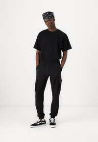 Svart bomulls T-skjorte og cargo-bukser med elastisk liv, sammen med svart og hvit sneakers, og et mønstret svart bandana.