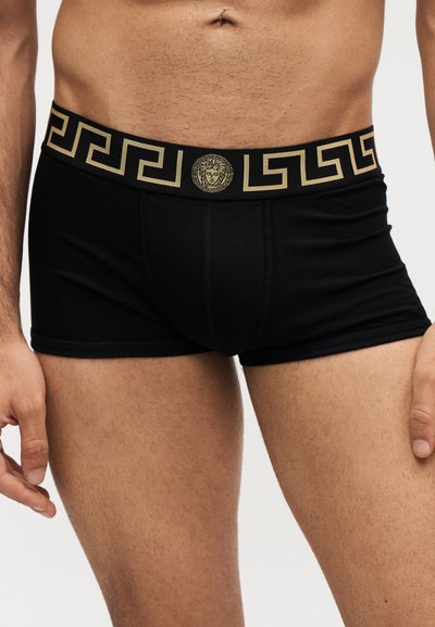 Homme portant un boxer noir avec une ceinture à motif doré et un logo tête de Méduse au centre devant.