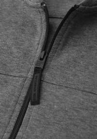 Grijze jas met rits, gemaakt van een gestructureerde stof, voorzien van een zwarte ritstreker met reliëfletters en contrasterende stiknaden langs de naden.