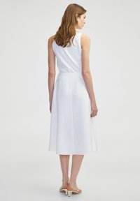 Weißes ärmelloses Kleid mit enger Wickeloptik und fließendem Rock. Hergestellt aus leichtem Stoff, mit feiner Textur und knielanger Länge.