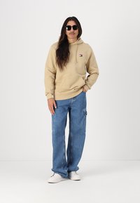 Beige hoodie with a front pocket and small logo, paired with loose blue jeans. White sneakers complete the outfit.
