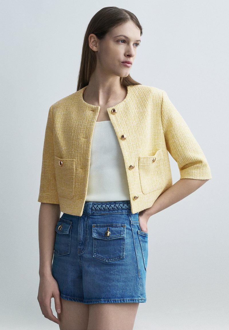 Jeune femme portant une veste jaune texturée à boutons, un haut blanc côtelé et un short en jean taille haute avec de grandes poches.