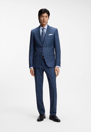 Mężczyzna w dopasowanym niebieskim garniturze z subtelnym kratkowanym wzorem, jasnoniebieskiej koszuli, wzorzystym niebieskim krawacie, białej poszetce i czarnych eleganckich butach.