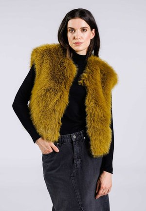 Gilet in pelliccia sintetica di colore giallo senape con una superficie morbida e testurizzata. Indossato sopra un dolcevita nero, abbinato a una gonna in denim scuro a vita alta.
