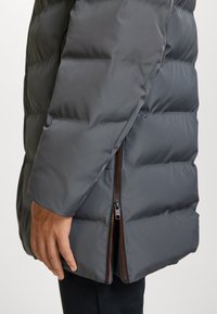 Giubbotto imbottito grigio scuro, con sezioni trapuntate, una cerniera con un dettaglio arancione e una texture di tessuto liscia e lucida.