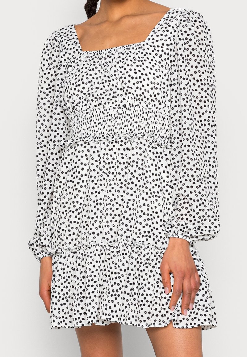 Vestido blanco con estampado floral negro, escote cuadrado, mangas largas abullonadas, cintura fruncida y dobladillo con volantes. Tela ligera y vaporosa.
