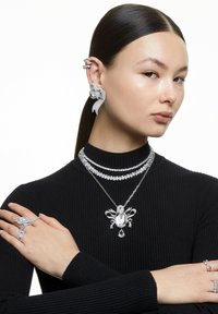 Frau mit glattem Haar, die ein schwarzes, geripptes Oberteil trägt, geschmückt mit funkelnden silbernen Statement-Ohrringen, mehrlagigen Halsketten und mehreren Ringen.