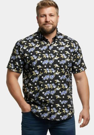 Uomo con barba che indossa una camicia nera a maniche corte con motivo floreale blu e bianco, in piedi con una mano in tasca.
