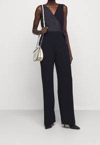 Merihenkinen haalari, jossa on ristiin menevä kaula-aukko ja ruutu-kuvio. Haalari on leveät lahkeet ja sitä on yhdistetty pieneen metalliseen laukkuun.